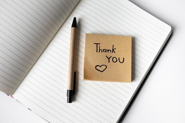 Sticky note with handwritten phrase thank you on a notebook with pen top view