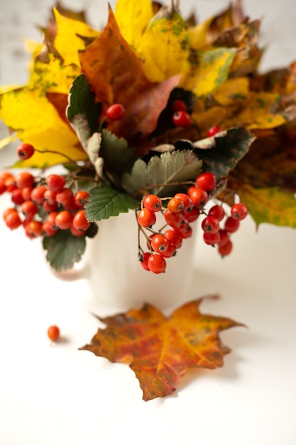 Still life. Autumn bouquet of bright fallen leaves and red rowan in a white cup on a wooden background. Vertical photo