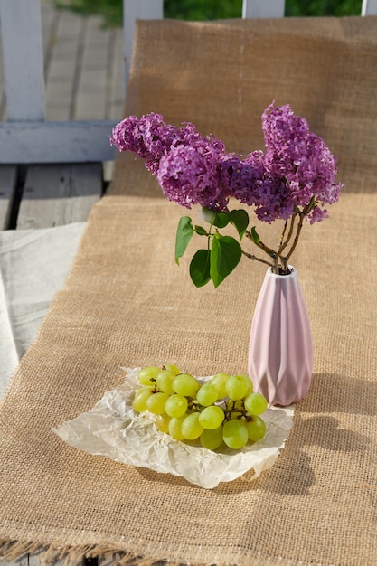 Still life and food photo A bunch of grapes and vase with lilacs stand on burlap on a wooden floor