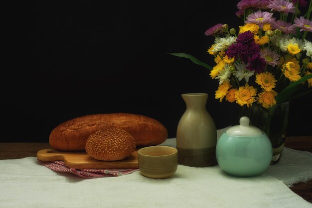 Still life of a tea set with breads placed on the wooden plate and flower bouquet in a glass.