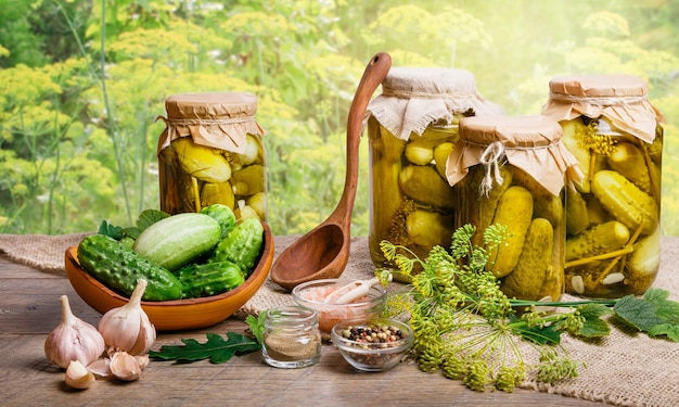 Still life with fresh and canned cucumbers in jars, spices on wooden table with sunlight blurred by a natural background. Preserved vegetables