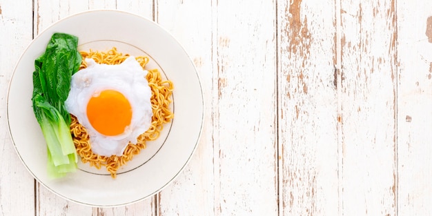 Stir fried spicy instant noodles with bok choy topped with fried egg in ceramic plate on white wood texture background with copy space for text, top view, flat lay, 2 x 1 ratio