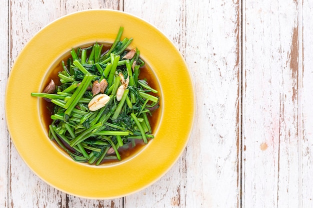 Stir fried water spinach with garlic in yellow ceramic plate on white old wood texture background with copy space for text, top view, light and airy food photography, morning glory