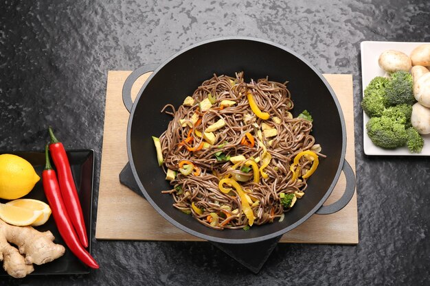 Photo stirfry tasty noodles with vegetables meat in wok and ingredients on dark textured table flat lay