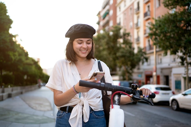 Stock photo of a beautiful short-haired woman using her phone on the street to unlock a public scooter. She's smiling.