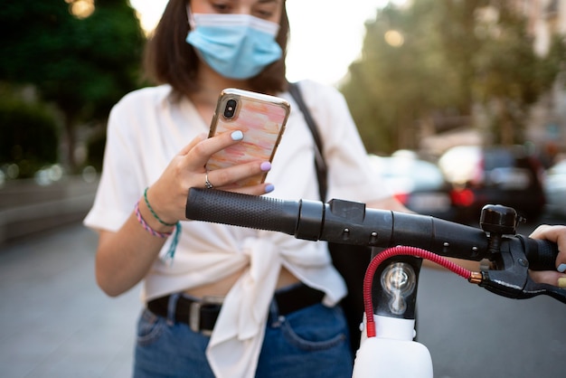 Stock photo of a beautiful short-haired woman using her phone on the street to unlock a public scooter. She's wearing a mask to protect herself.