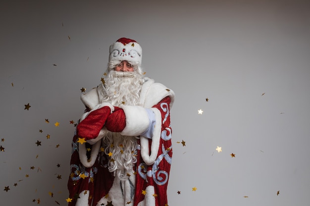 Stock photo of jolly Father Frost in festive clothing with long white beard outstretching his arms and smiling at camera under flying golden confetti. Isolated on grey background.