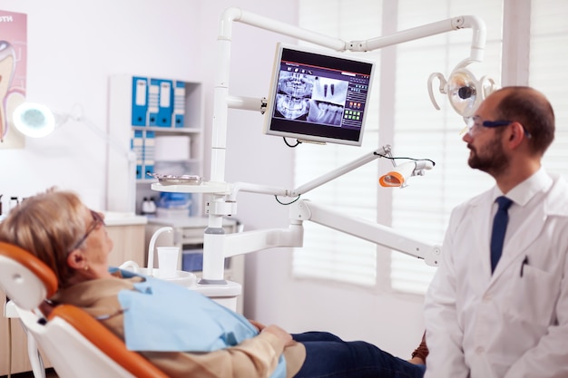 Photo stomatolog talking about teeh treatment to senior woman in dentist cabient. medical teeth care taker pointing at patient radiography on screen sitting on chair.