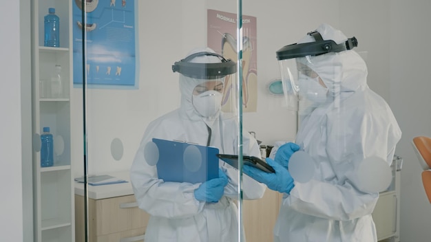 Stomatologists wearing hazmat suits using tablet for dental hygiene and consultation. Dentist and assistant doing patient examination with professional equipment during pandemic