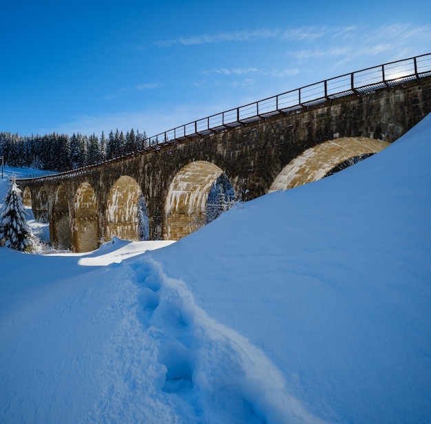 Stone viaduct arch bridge on railway through mountain snowy fir forest Snow drifts on wayside and hoarfrost on trees