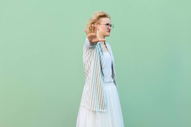 Photo stop it. portrait of serious young blonde woman in white shirt, skirt, and striped blouse with eyeglasses standing with stop hand gesture and blocking. studio shot isolated on light green background.