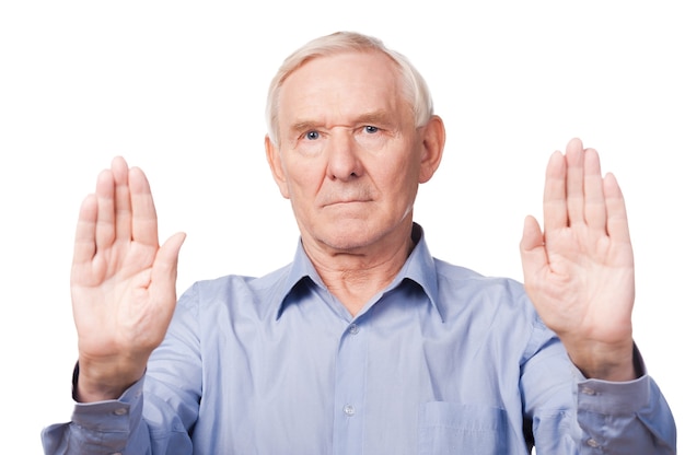 Stop it! Serious senior man in shirt showing his palms and looking at camera while standing against white background