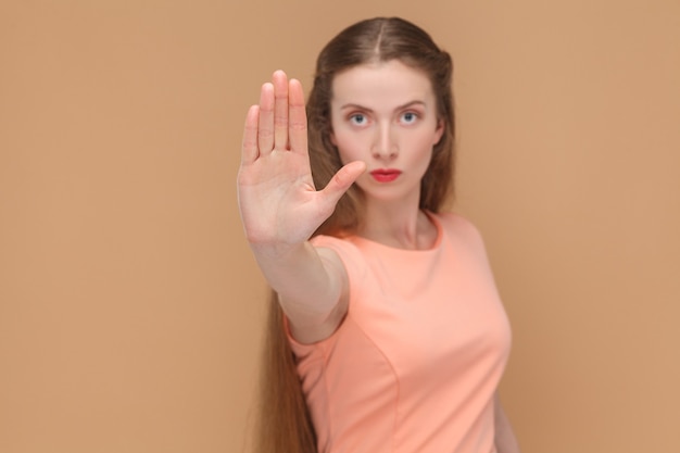 Stop it. woman with ban sign looking at camera. portrait of emotional cute, beautiful woman with makeup and long hair in pink dress. indoor, studio shot, isolated on light brown or beige background.