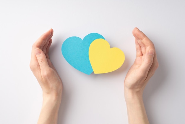 Stop the war in Ukraine concept First person top view photo of female hands protecting two yellow and blue hearts on white background