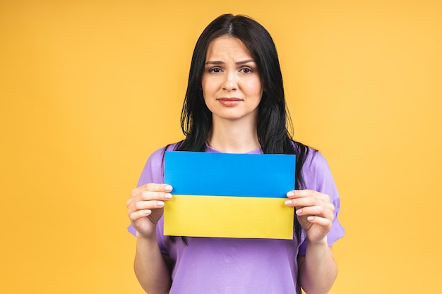 Stop war in Ukraine Portrait of sad agressive angry woman holding in hands Ukrainian flag isolated over yellow background