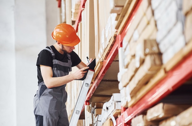 Storage worker stands on the ladder in uniform and notepad in hands and checks production.