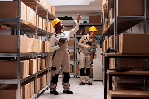 Storehouse manager checking products stock supply in shop logistics department warehouse. African american woman using inventory application on tablet and reaching for package on shelf