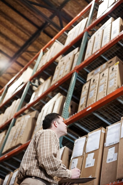 A storeroom manager looking at stock stacked high on shelves making notes with a clipboard and pen