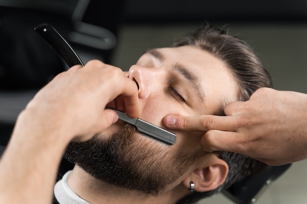 Straight razor cut mans beard in barbershop. Barber man making hairstyle for handsome man.