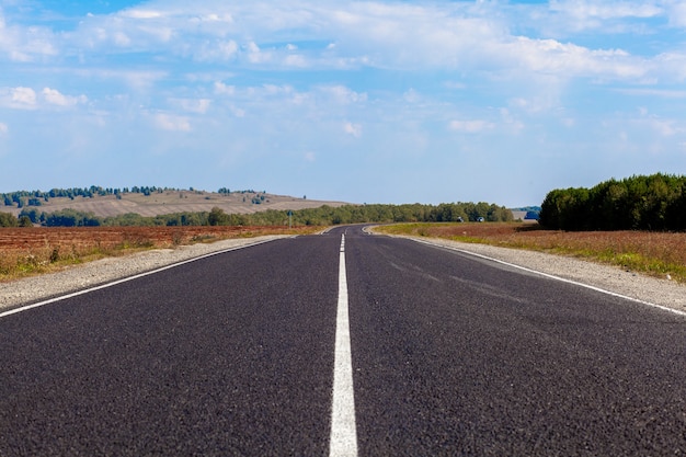 Straight road with a marking on the nature background. Open Road in future, no cars, auto on asphalt road through green forest, trees. Clouds on blue sky in summer, sunshine, sunny day. Bottom view