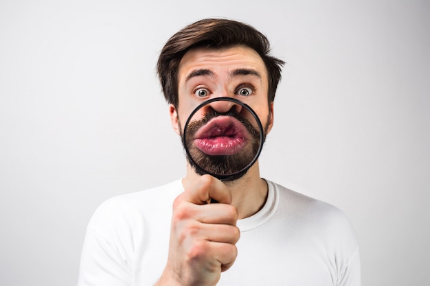 Strange and amazed man isolated on white wall and making some fun with putting a loupe in front of his mouth. Guy looks quite unusual from this angle. Close up. Cut view