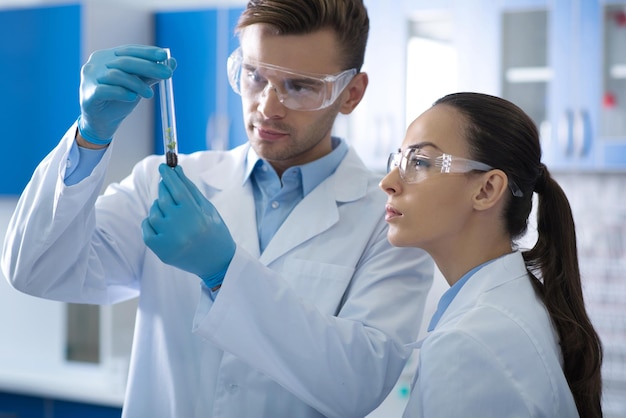 Strange case Young serious responsible scientists in a glasses and rubber gloves standing in the laboratory focusing at the vial and looking like interested