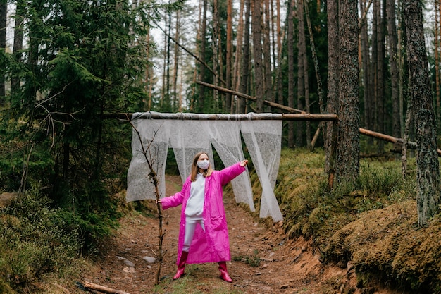 Strange young woman in mask and pink raincoat posing with stick in the forest