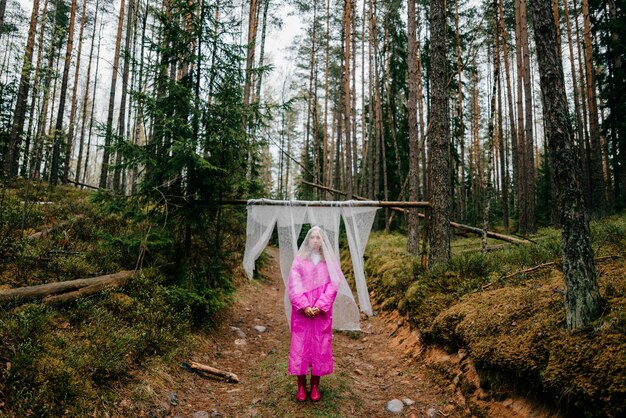 Strange young woman in a pink raincoat posing with covering cheesecloth in the forest