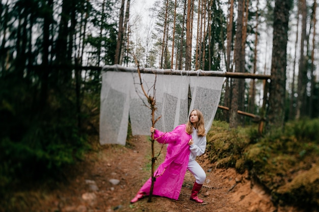 Strange young woman in pink raincoat posing with stick in the forest