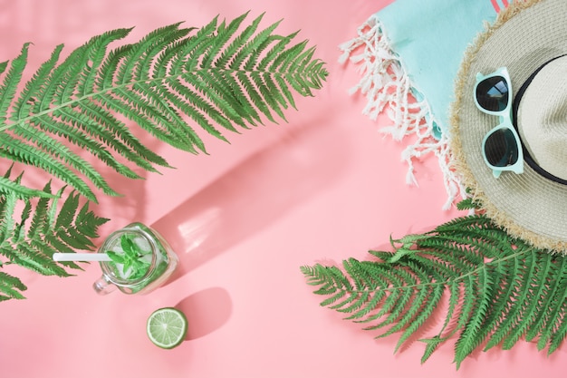 Straw beach hat, sunglasses, towel, leaf of fern and refreshing drinks with lime on pastel pink