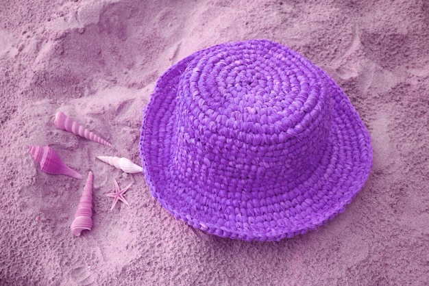 Straw hat with many types of little seashells on the sandy beach in pastel purple color tone