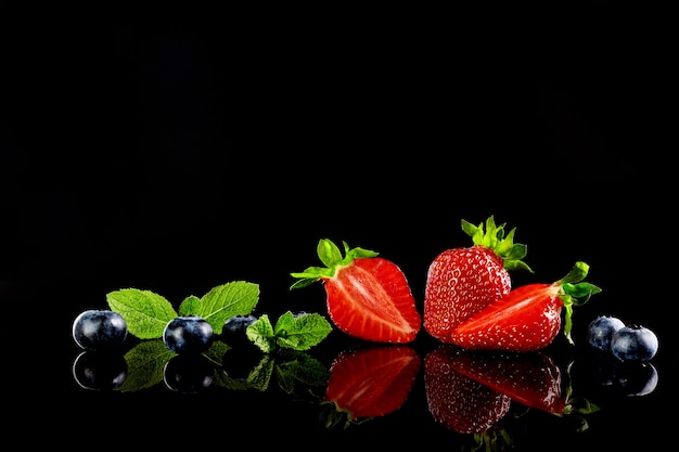 Strawberries and blueberries with mint leafs on the black glass background with a reflection. Selective focus.