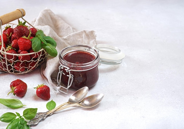 Strawberry jam with basil in the glass jar and a basket with berries at white table copy space