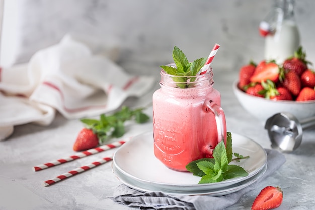 Strawberry and milk or yogurt smoothies in a beautiful red glass jar and sprigs of mint on a white concrete table.