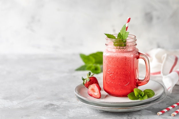 Strawberry and milk or yogurt smoothies in a beautiful red glass jar and sprigs of mint on a white concrete table.