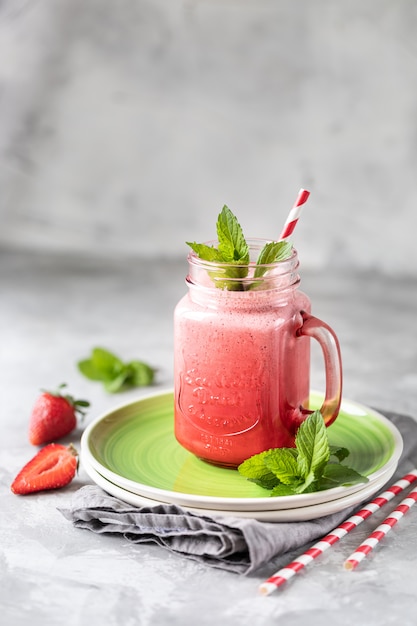 Strawberry and milk or yogurt smoothies in a beautiful red glass jar and sprigs of mint on a white concrete table.