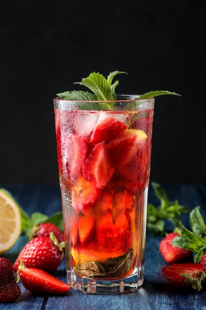 Strawberry Mojito. Cold summer mojito cocktail with strawberries, mint, lemon and ice in a glass on a blue wooden table. on a dark background