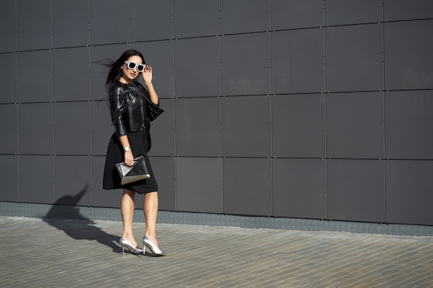 Street fashion look. Beautiful young woman in stylish sunglasses wearing elegant dress and fashionable black fringed leather jacket holding clutch in the hands.