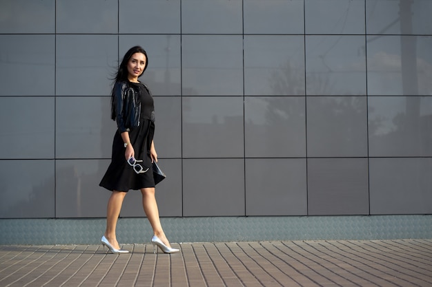 Street fashion look. Pretty young woman wearing elegant black dress and fashionable black fringed leather jacket holding stylish sunglasses in the hand.  Urban wall on  Copy space.