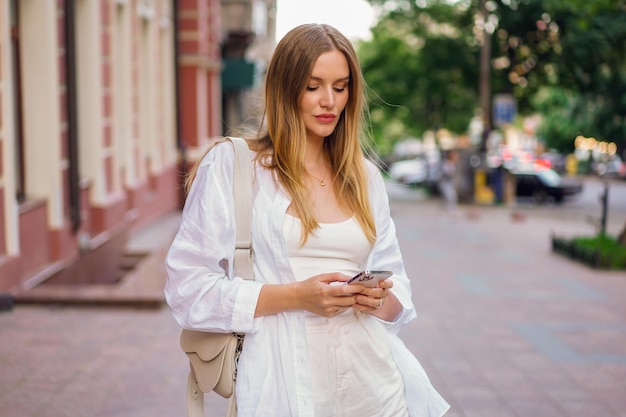 Street fashion portrait of stylish woman wearing white spring summer outfit tap on her smartphone, posing on european city.