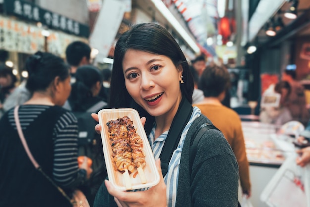 Street food grilled chicken barbecue on plate young girl traveler showing to camera standing at vending stand with teeming crowd background. tourist smiling sharing delicious yakitori japanese local.