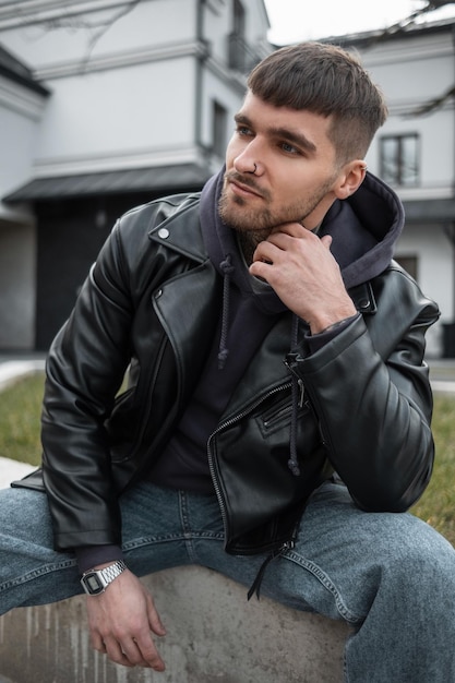 Street portrait of a handsome fashionable hipster man with a hairstyle and beard in stylish casual clothing with a leather jacket and hoodie sitting and poses on the street