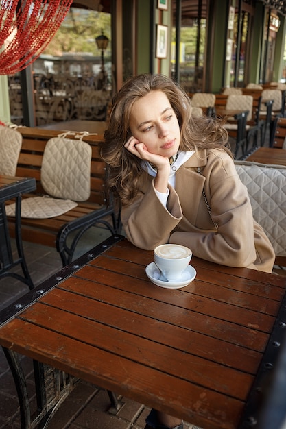 Street portrait of young woman wearing beige coat drinking coffee on cafe veranda with dreamy and thoughtful gaze