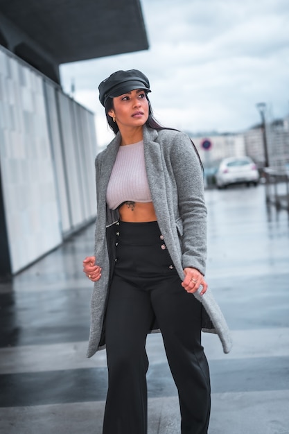 Street style in the city of a brunette Latin girl with a leather hat and a gray raincoat walking in autumn