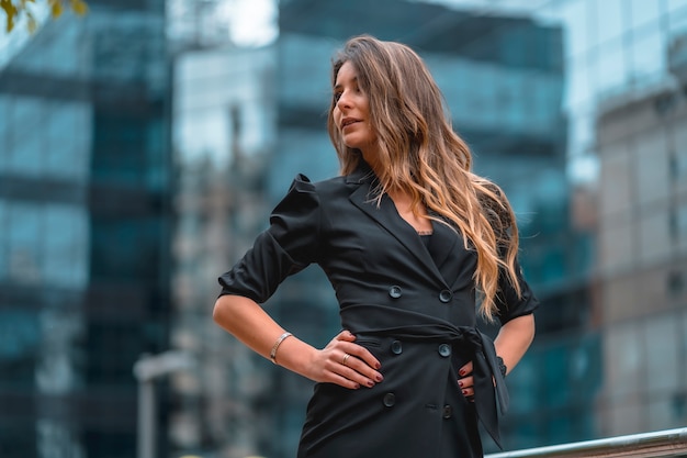 Street style, an enterprising young blonde Caucasian woman in a black suit in the black glass building where she works in the background.