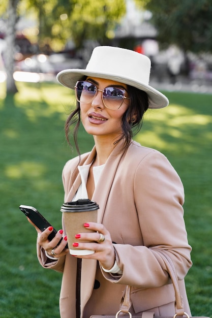 Street style portrait of blogger with smartphone and coffee cup. Girl weared white hat, dress and jacket.