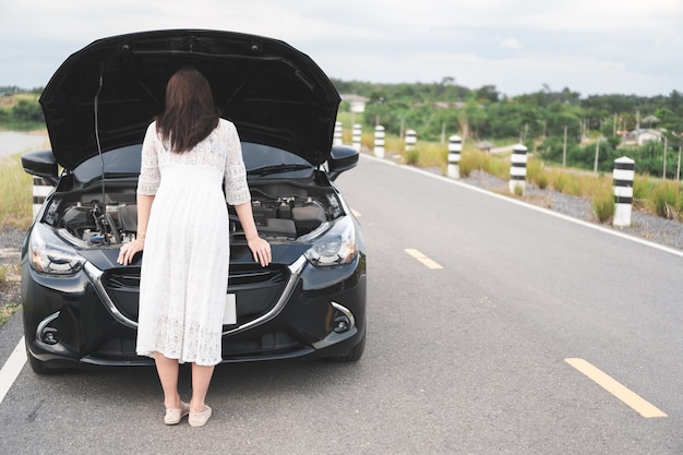 Stressed asian woman open hood, looking a broken car and checking engine on country road.