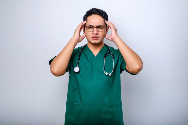 Stressed Asian young doctor or male nurse in green uniform with stethoscope having headache