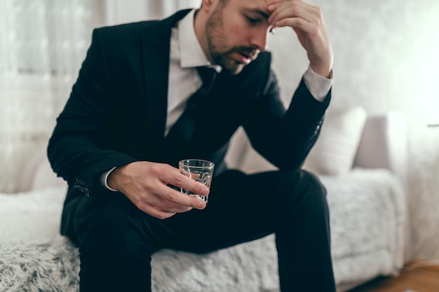 Stressed businessman sitting on the couch in suit, drinking alcohol and holding hand on head.