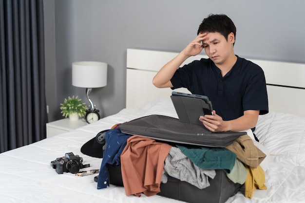 Stressed man using tablet and packing clothes into suitcase on a bed holiday travel concept